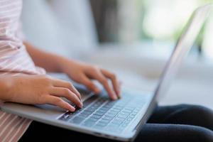 garotinha asiática da escola usa computador portátil sentado no sofá sozinho em casa. criança aprendendo lendo conteúdo de mídia social on-line, jogando lições de educação conversando com amigos. foto