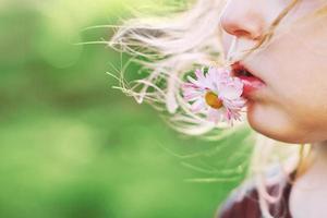 menina com uma margarida flor dentro dela boca em uma verde fundo fechar. lábios com flor em Relva fundo. retrato do uma pequeno menina