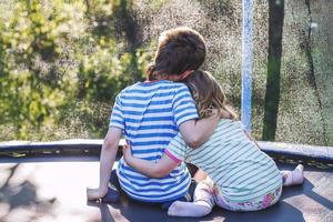 crianças abraçando dentro a jardim em trampolim. irmão com dele pequeno irmã ao ar livre costas foto