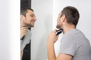 uma homem faz a barba dele barba com uma aparador , parece dentro a espelho e sorrisos foto