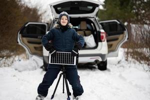 Garoto com solar painel bateria em tripé contra carro dentro inverno madeiras. foto