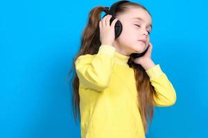 retrato do uma menina em uma azul fundo dentro uma amarelo suéter, uma pequeno menina com sem fio fones de ouvido. foto