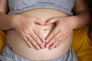 foto do feliz ásia grávida mulher sentado dentro cama e tocante dela barriga às casa com pequeno sapatos para a por nascer bebê dentro a barriga