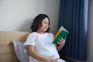 jovem ásia grávida mulher lendo livro em a cama às casa enquanto segurando dela barriga foto