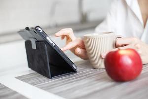 mulher com copo do café ou chá usando computador portátil dentro quarentena bloqueio dentro a cozinha dentro branco camisa. menina com uma tábua foto