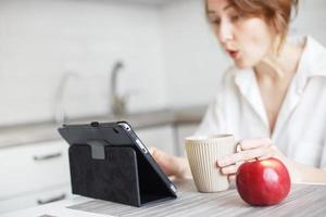 feliz surpreso mulher com copo do café ou chá usando computador portátil dentro a cozinha dentro branco camisa. menina com uma tábua foto