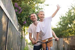 uma pai passeios dele filho dentro uma branco camiseta em uma bicicleta. a feliz filho espalhar dele braços foto