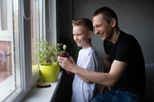 uma fofa Garoto é rega interior plantas. Papai Próximo para dele filho perto a janela foto