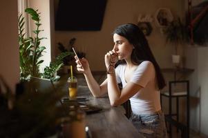 a menina é sentado dentro uma cafeteria às a bar, olhando às a telefone, segurando óculos dentro dela mão, uma lapto foto