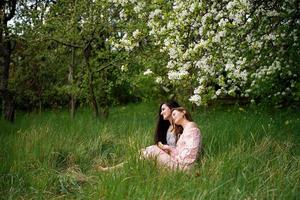dois jovem meninas dentro vestidos estão sentado em a verde Relva debaixo uma branco árvore e rindo foto