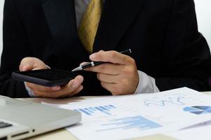 a mão do empresário segurando um telefone celular e uma caneta na mesa tem documentos comerciais, gráficos e relatórios financeiros foto