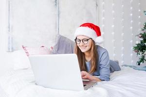 feliz menina dentro santa chapéu compras conectados em computador portátil e sentado em cama Próximo foto