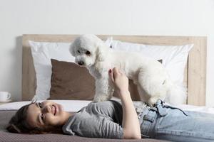 lindo menina às casa com uma cachorro. a anfitriã com uma bichon tocam dentro cama. foto