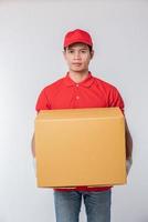 imagem de um jovem entregador feliz em uniforme de camiseta em branco de boné vermelho em pé com caixa de papelão marrom vazia isolada no estúdio de fundo cinza claro foto