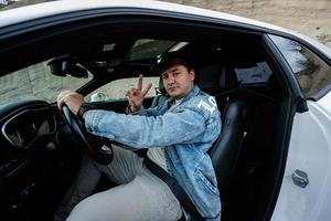 bonito homem dentro jeans Jaqueta e boné sentar às dele branco músculo carro e mostrar dois dedos. foto