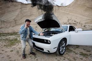 homem chocado chamando phoen para emergência perto de seu muscle car quebrado, capô aberto, fumaça do motor, avaria, precisa de ajuda. foto