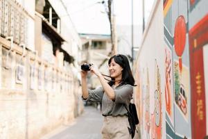 jovem ásia mulher mochila viajante usando digital compactar Câmera, desfrutando rua cultural local Lugar, colocar e sorriso. viajante verificação Fora lado ruas. foto