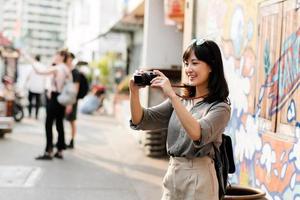 jovem ásia mulher mochila viajante usando digital compactar Câmera, desfrutando rua cultural local Lugar, colocar e sorriso. viajante verificação Fora lado ruas. foto