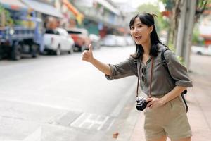 sorridente jovem ásia mulher viajante pedindo carona em uma estrada dentro a cidade. vida é uma viagem conceito. foto