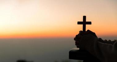 silhueta do jovem mulher ajoelhado baixa Rezar e segurando cristão Cruz para adorando Deus às pôr do sol fundo. conceito do cristão ajoelhado e Rezar para Deus. foto