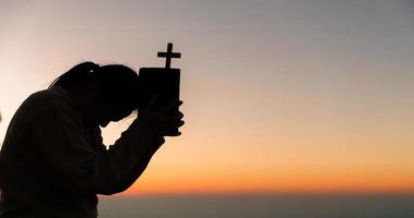 silhueta do jovem mulher ajoelhado baixa Rezar e segurando cristão Cruz para adorando Deus às pôr do sol fundo. conceito do cristão ajoelhado e Rezar para Deus. foto