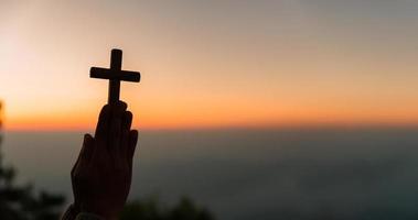 silhueta do jovem mulher ajoelhado baixa Rezar e segurando cristão Cruz para adorando Deus às pôr do sol fundo. conceito do cristão ajoelhado e Rezar para Deus. foto