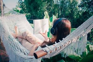 jovem relaxado mulher ouvindo para música com fones de ouvido lendo livro em branco maca dentro jardim às lar. manhã Sol. lento vivendo, final de semana lazer atividade. quarentena e auto isolamento período. foto