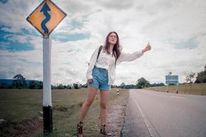 lindo jovem ásia mulher com a mochila em pé perto uma tráfego placa e espera em a asfalto estrada dentro quente clima, uma menina para pegar uma passagem carro para viajando. Mochileiro mostrando uma mão gesto. foto