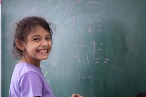 feliz estudante retrato do pequeno menina escrevendo em verde quadro-negro dentro sala de aula, Educação e escola conceito foto