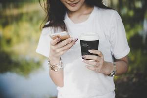 jovem segurando uma xícara de café descartável enquanto envia mensagens de texto pelo smartphone ao ar livre foto