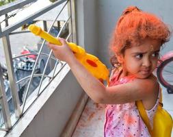 doce pequeno indiano menina jogando cores em holi festival, segurando pichakaree cheio do cores, holi festival celebrações dentro Délhi, Índia foto