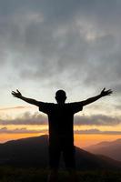 silhueta do jovem homem em pé em topo do montanha pico com dele mãos elevado acima dele cabeça Como símbolo do adoração e invocação do Deuses bênçãos com crença e poder do fé dentro Deus. foto