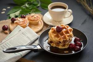 waffles e cerejas com mel em uma panela, tortas de amêndoas crocantes e uma xícara de café na mesa preta