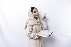 jovem ásia muçulmano mulher sorridente enquanto apontando para cópia de espaço ao lado dela e segurando a Alcorão foto