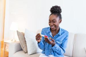 jovem mulher olhando às gravidez teste dentro felicidade. finalmente grávida. atraente Preto mulheres olhando às gravidez teste e sorridente enquanto sentado em a sofá às casa foto