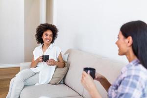 dois fêmea amigos dentro social distanciar sentado em sofá. melhor amigos tendo café juntos enquanto separado de social distanciar em sofá às casa foto