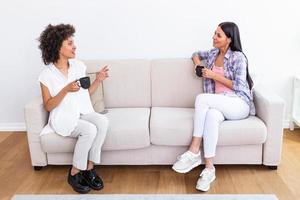 dois fêmea amigos dentro social distanciar sentado em sofá dentro coronavírus pandemia tempo. melhor amigos tendo café juntos enquanto separado de social distanciar em sofá às casa foto