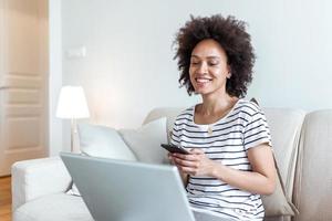 feliz jovem Preto mulher usando Móvel telefone enquanto sentado uma sofá às casa com computador portátil computador. sorridente mulher sentado em sofá relaxante enquanto navegando conectados compras local na rede Internet. foto