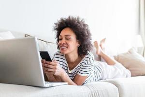 feliz jovem Preto mulher usando Móvel telefone enquanto sentado uma sofá às casa com computador portátil computador. sorridente mulher sentado em sofá relaxante enquanto navegando conectados compras local na rede Internet. foto