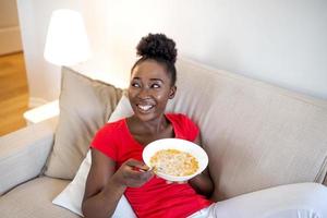 sorridente mulher comendo saudável café da manhã às casa dentro manhã, relaxante em sofá dentro vivo sala. lindo feliz africano americano menina desfrutando Comida dentro tigela sentado em sofá foto