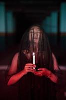 a ásia mulher com fechadas olhos e uma transparente véu manchado com sangue detém uma vela dentro dela mão durante satânico adoração foto