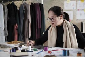 designer de moda feminina asiática de meia-idade trabalha em estúdio por meio de esboços de desenho de ideias com tablet digital e tecido colorido para uma coleção de design de vestidos, alfaiate profissional e empresário de sme. foto