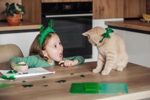 uma pequeno menina com uma aro decorado com trevo desenha e cortes verde trevos para st. patrick's dia às a mesa às casa dentro a cozinha, Próximo para dela é dela lindo gato foto