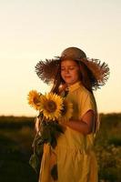 uma feliz jovem menina com grandes cabelo dentro uma Palha chapéu carrinhos dentro uma ampla campo do girassóis. verão dia. uma caloroso pôr do sol. foto