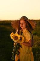 uma feliz jovem menina com grandes cabelo dentro uma Palha chapéu carrinhos dentro uma ampla campo do girassóis. verão dia. uma caloroso pôr do sol. foto