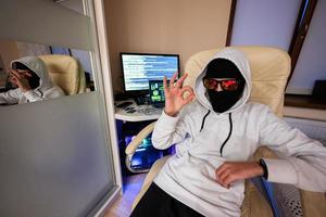 Garoto hacker mostra Está bem placa. Internet roubo . homem vestindo uma balaclava Sentou atrás uma computador portátil. foto