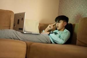 jovem adolescente Garoto dentro frente do uma computador portátil em uma cama às tarde. foto