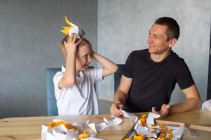 uma fofa Garoto é sentado com dele Papai às a mesa e colecionar origami. de papagaio cabeça fez do papel foto