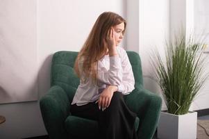 uma jovem menina dentro uma branco camisa é sentado dentro uma cadeira dentro a do psicólogo escritório foto