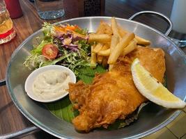 fechar acima Visão do delicioso peixe e salgadinhos com francês fritas, César salada com balsâmico molho e tártaro molho ou maionese foto